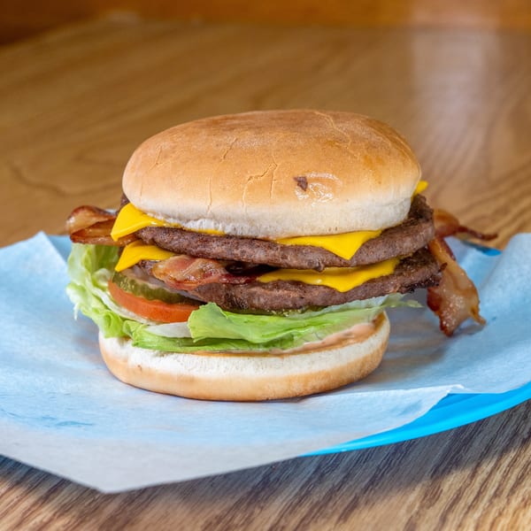 Double cheeseburger with bacon, lettuce, tomato, pickles, and sauce on a bun, served on blue-lined paper over a wooden table.
