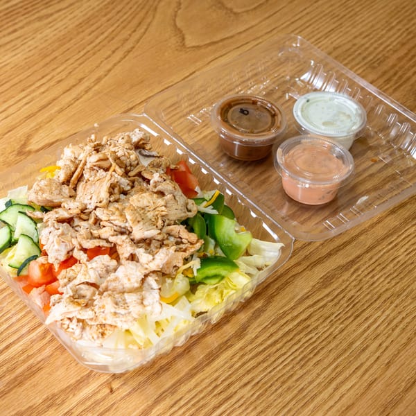 Takeout container of grilled chicken salad with lettuce, cucumbers, tomatoes, peppers, and three dipping sauces on a wooden table.