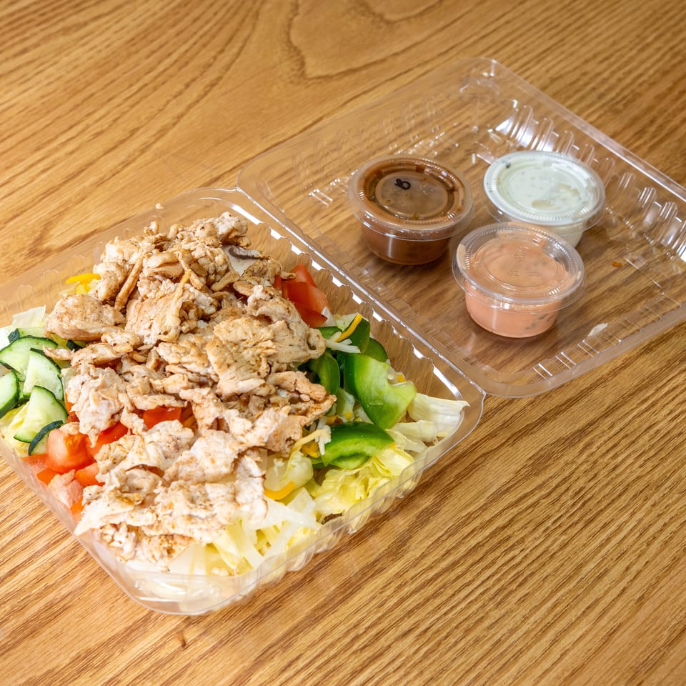 Takeout container of grilled chicken salad with lettuce, cucumbers, tomatoes, peppers, and three dipping sauces on a wooden table.